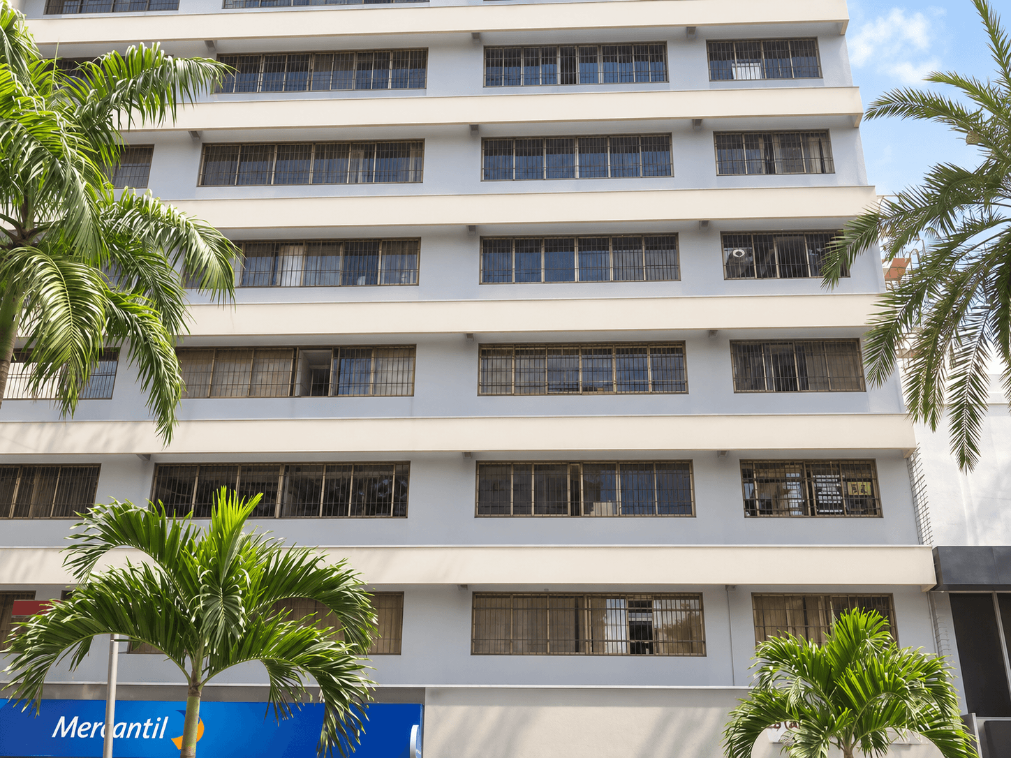 Fachada del Edificio Torre Royal: seis pisos de ventanas alargadas sobre una fachada blanca, con el letrero azul del Banco Mercantil en la planta baja y palmeras delante